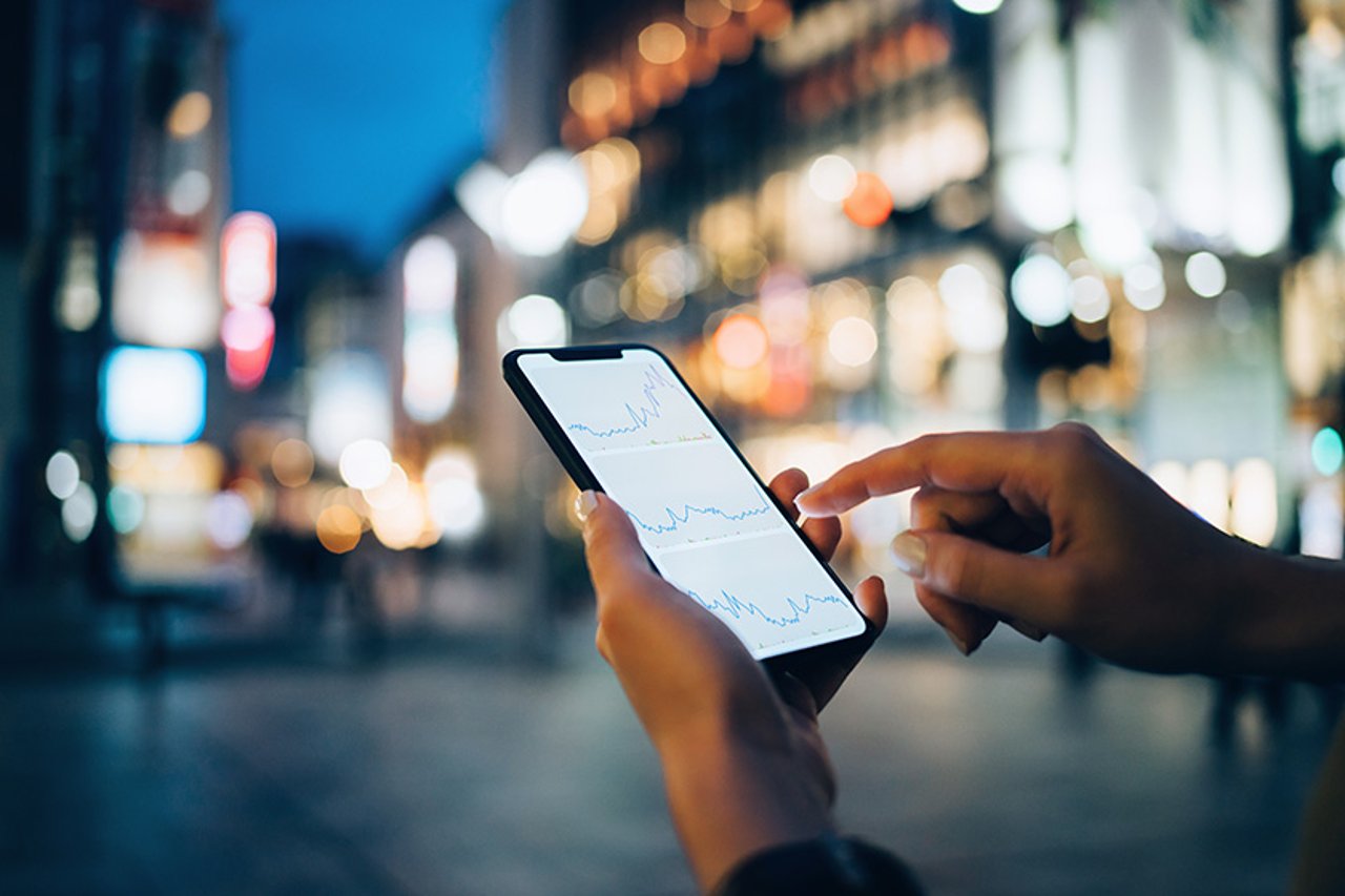 Las compañías de servicios financieros innovadoras están utilizando API de comunicaciones para transformar la experiencia del cliente, deleitando a los clientes, a veces indecisos, con interacciones de autenticación en tiempo real, vídeo, voz y mensajería. Businesswoman reading financial trading data on smartphone in downtown city street against illuminated urban skyscrapers